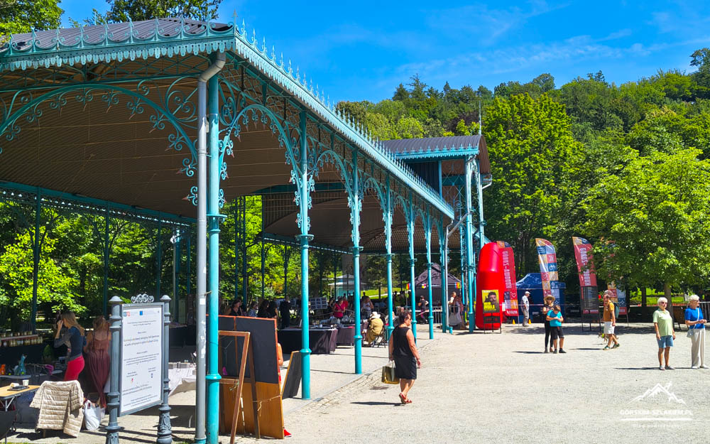 Decorative blue iron pavilion in a sunny park with stalls, banners, and people walking around the event area.