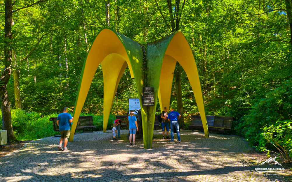 Yellow curved arch sculpture in a forested park with visitors nearby and benches nearby nearby trees, sunlight filtering through leaves.