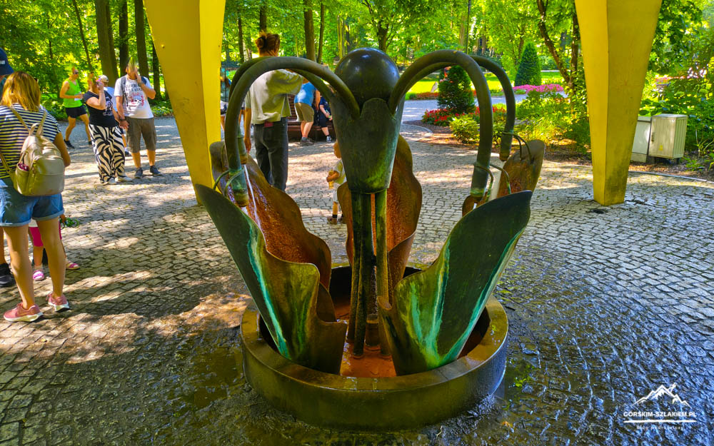 Metal sculpture fountain in a park surrounded by visitors and trees with sunlight filtering through the leaves. Bromze-green petals form a circular basin.