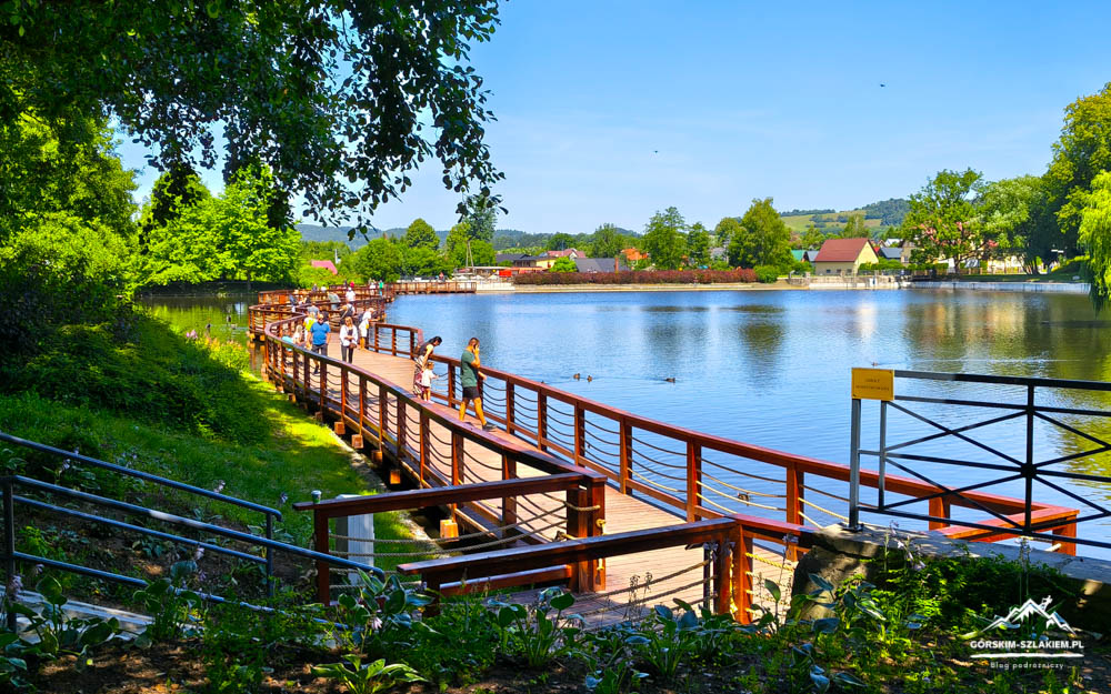 Wooden boardwalk over a calm lake with people strolling, surrounded by green trees and a distant village on a sunny day.