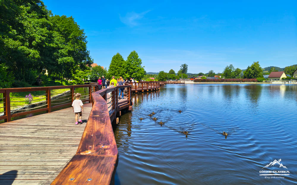 Wooden boardwalk along a blue lake with people strolling, a child walking near railing, ducks on the water, green trees and houses in the distance, sunny day.