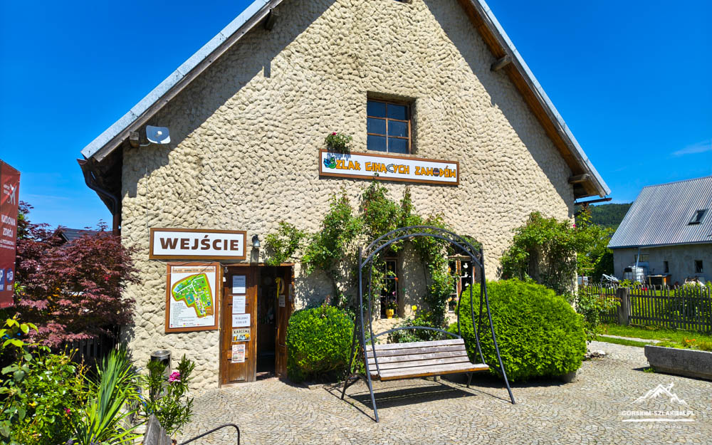 Stone cottage with an open wooden door marked 'WEJŚCIE', and a metal arch swing with surrounding greenery in a sunny courtyard.