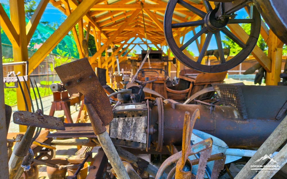 Rusty vintage machinery under a wooden shelter, with a large wheel visible in the upper right corner.