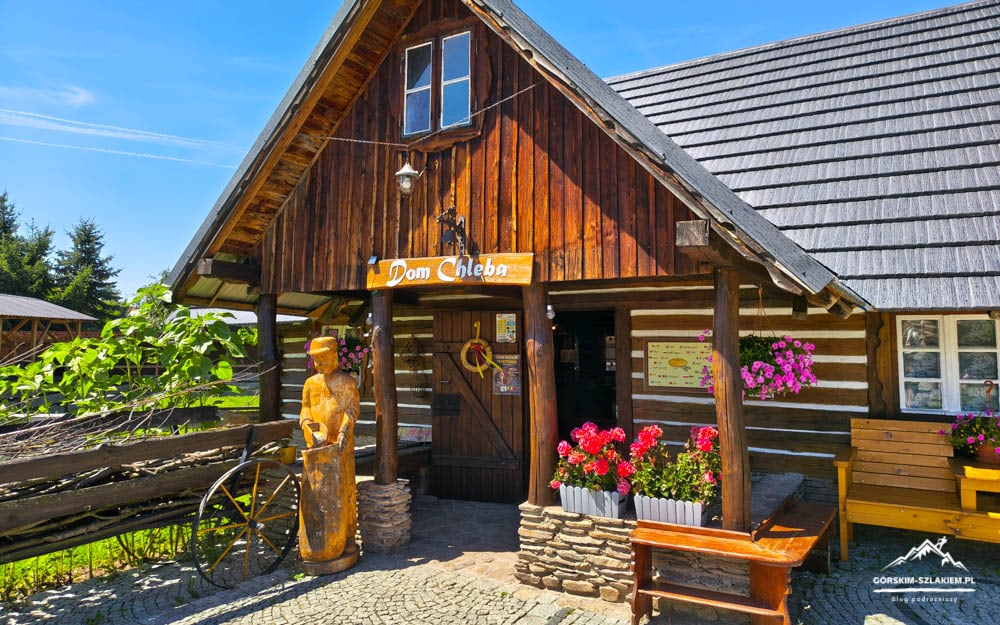 Rustic wooden bakery with a steep roof, flower boxes along the entrance, and a carved wooden statue beside a wagon wheel.