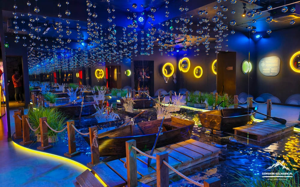 Indoor water-installation with small wooden boats on a shallow pool, neon blue lighting, and a ceiling of hanging glass spheres.