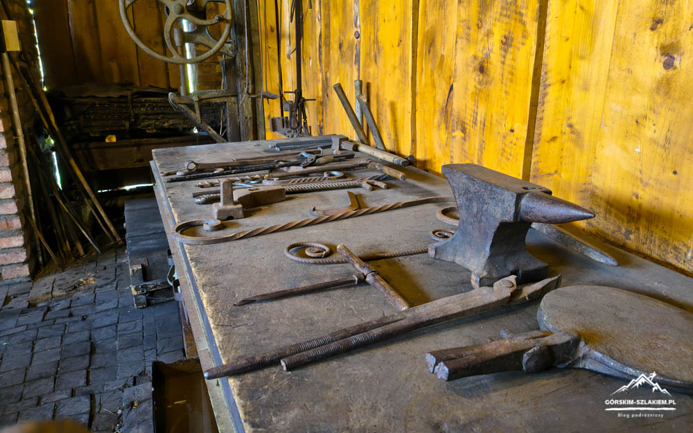 Blacksmith shop bench with an anvil and scattered hand tools in a rustic workshop against a yellow wooden wall.