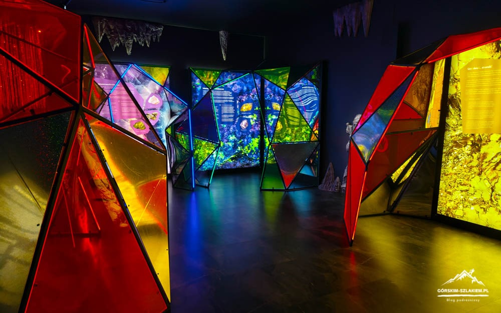 Colorful geometric glass sculptures forming a tunnel in a dark gallery, illuminated by neon lights.