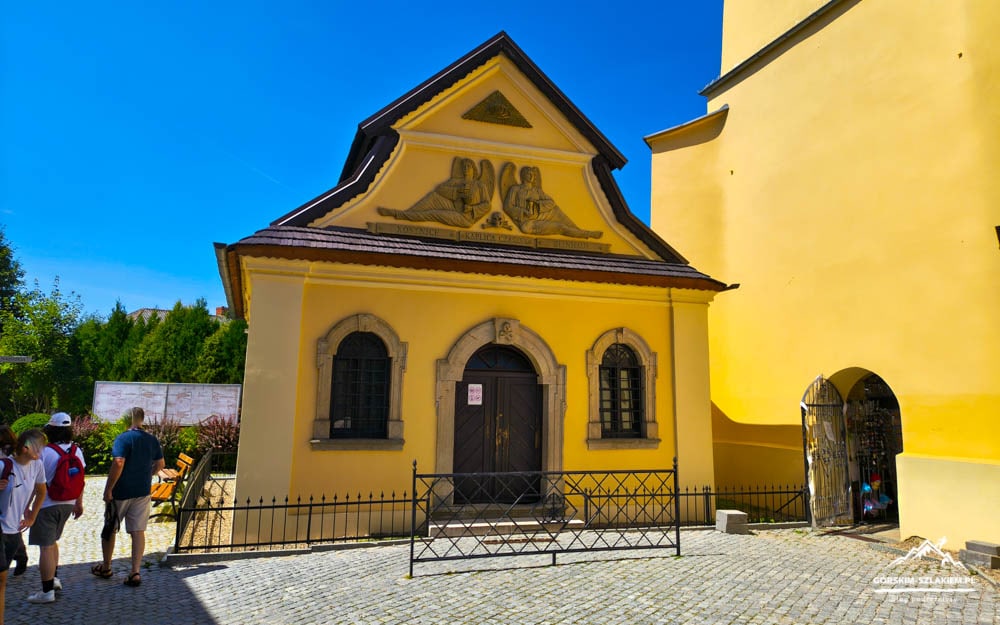 Small yellow chapel with a decorative gable relief and dark wooden door, set in a cobblestone square under a clear blue sky, with visitors on the left.