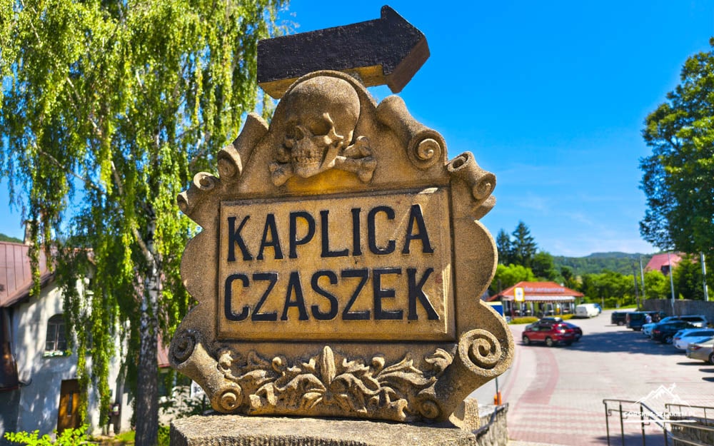 Stone sign reading 'Kaplica Czaszek' with a skull motif, beside a parking lot on a sunny day.