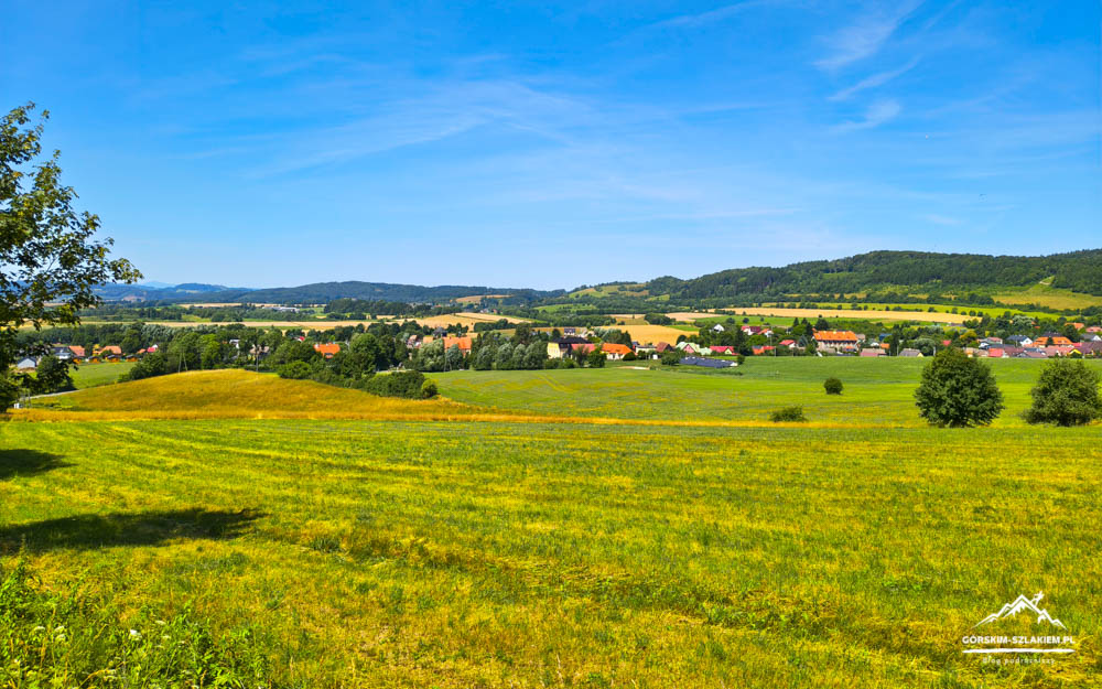 Wide countryside with golden and green fields, a small village of red-roofed houses in the distance, and tree-covered hills under a bright blue sky. (logo bottom-right)