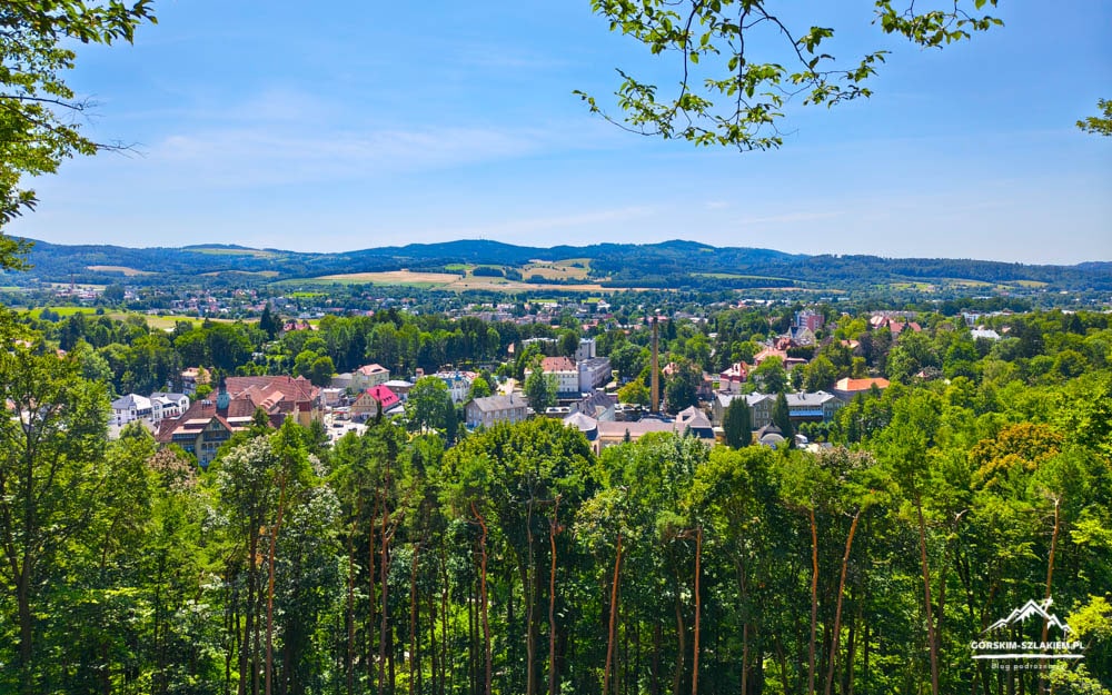 Panoramic view of a town in a valley, framed by a dense forest and distant green hills under a clear blue sky, from a hilltop.