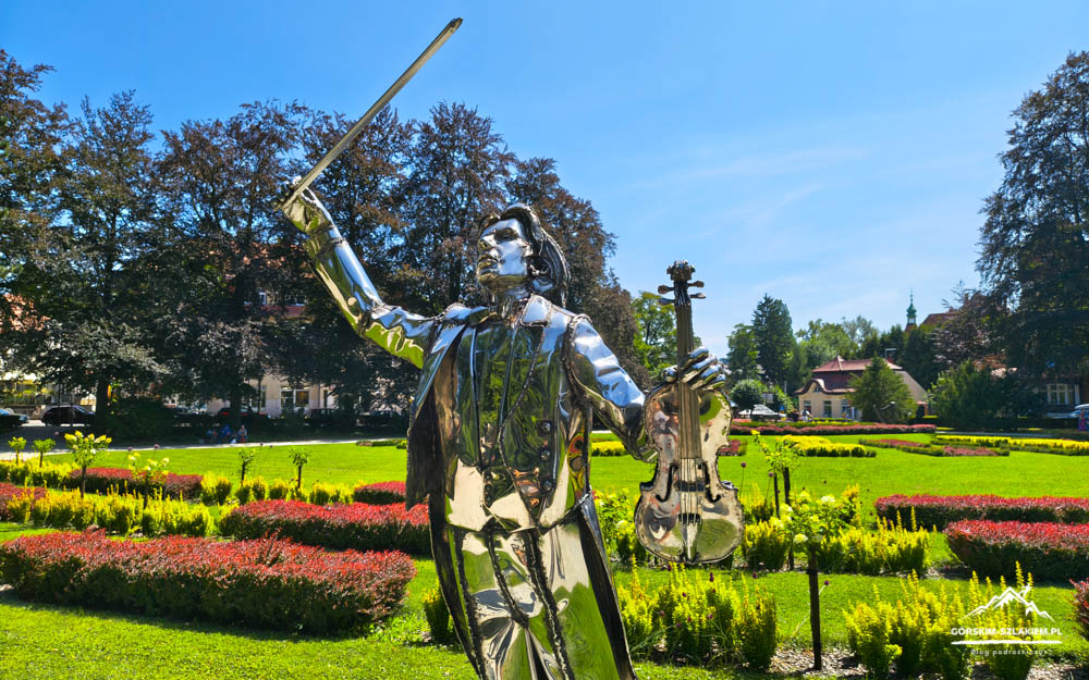 Shiny metal statue of a guitarist raising a sword in a park, with a reflective surface and a violin tucked under one arm, surrounded by colorful flower beds.