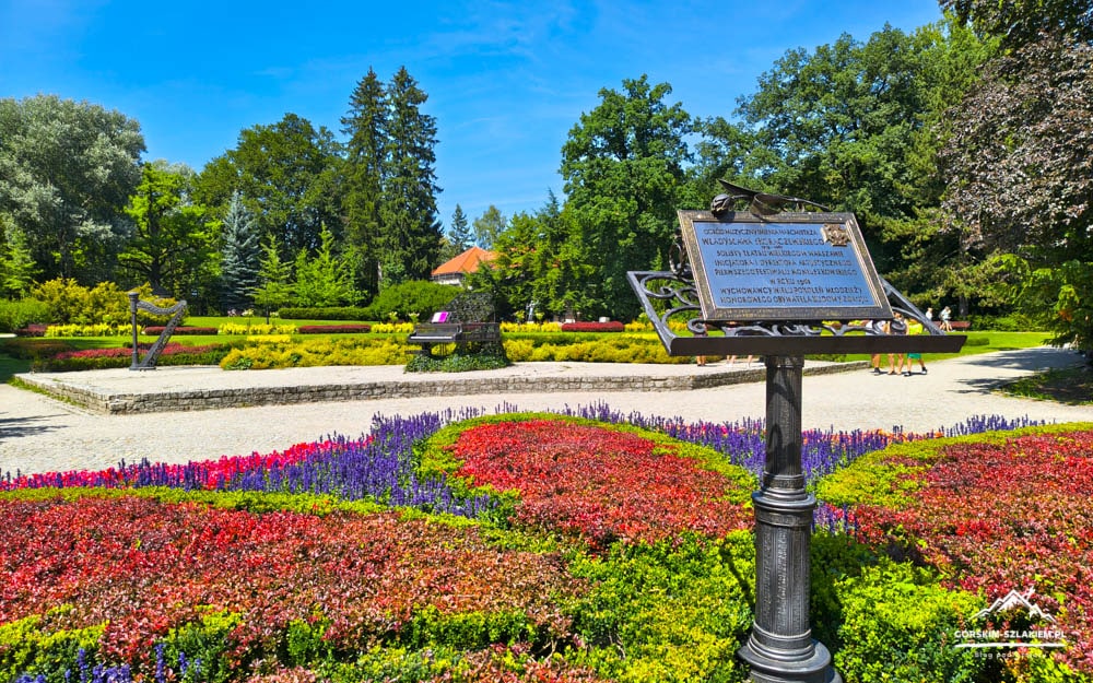 Sunlit park garden with multicolored flower beds, a metal plaque on a stand in the foreground, and a gravel path under a clear blue sky.