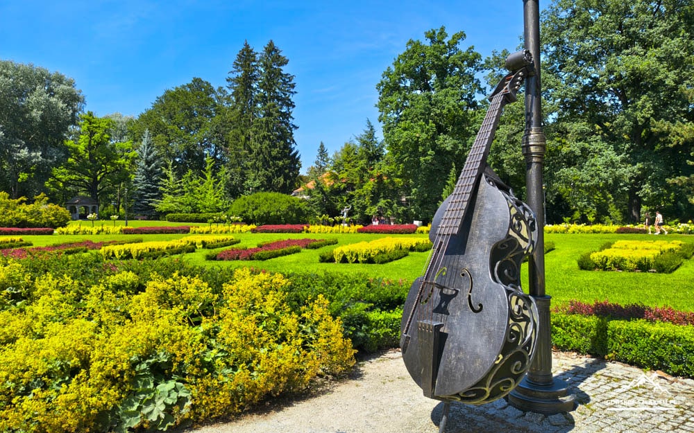 Metal guitar sculpture anchored to a lamppost in a sunlit public garden with colorful flower beds and trimmed hedges.