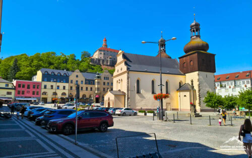 Bright town square with pastel buildings, a beige church, and a tall wooden bell tower under a blue sky.