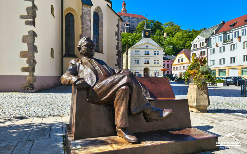 Bronze statue of a man seated in a chair in a sunlit cobblestone square with a church wall and pastel town buildings behind.