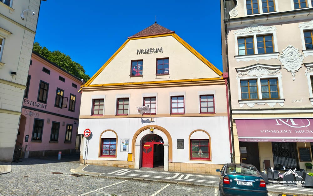 Front view of a small museum building with yellow trim and red doorway, labeled MUZEUM, on a sunny cobblestone street.