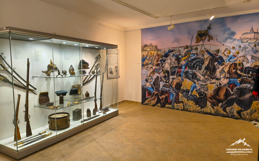 Museum gallery with a glass display case of antique weapons on the left and a large battle mural on the right wall.