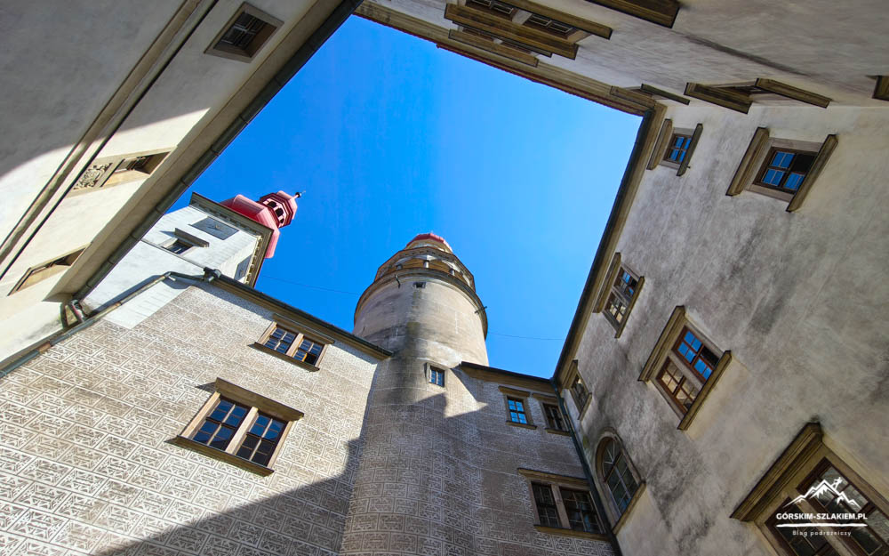 Viewed from a narrow courtyard up toward a tall historic tower against a bright blue sky; surrounding beige buildings with windows frame the scene.