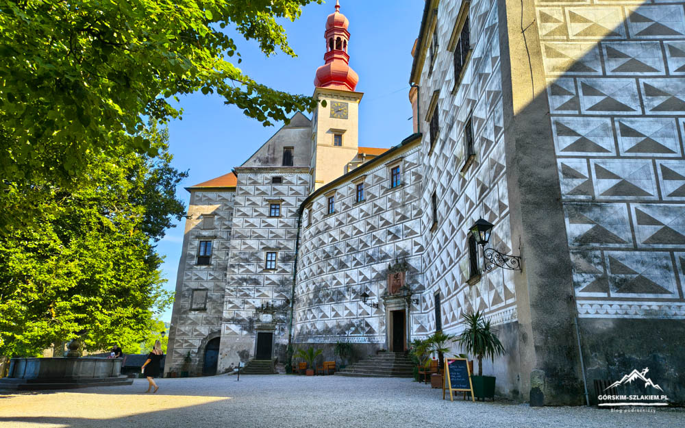 Castle courtyard with white patterned walls, a red-topped tower, and lush green trees on the left; a chalkboard sign sits near the entrance.