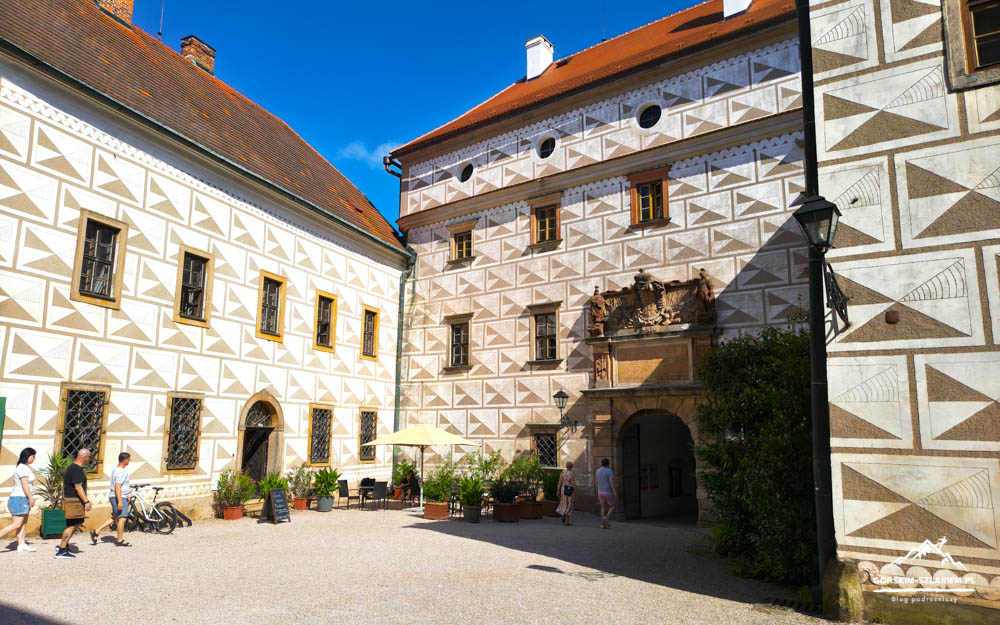 Sunlit European courtyard with white patterned walls, an arched doorway, and outdoor seating under a yellow umbrella.