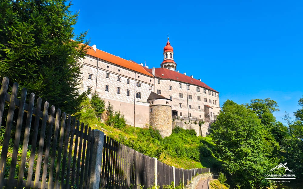 Castle atop a hill with red-tiled roofs and stone walls, seen beyond a wooden fence against a bright blue sky.