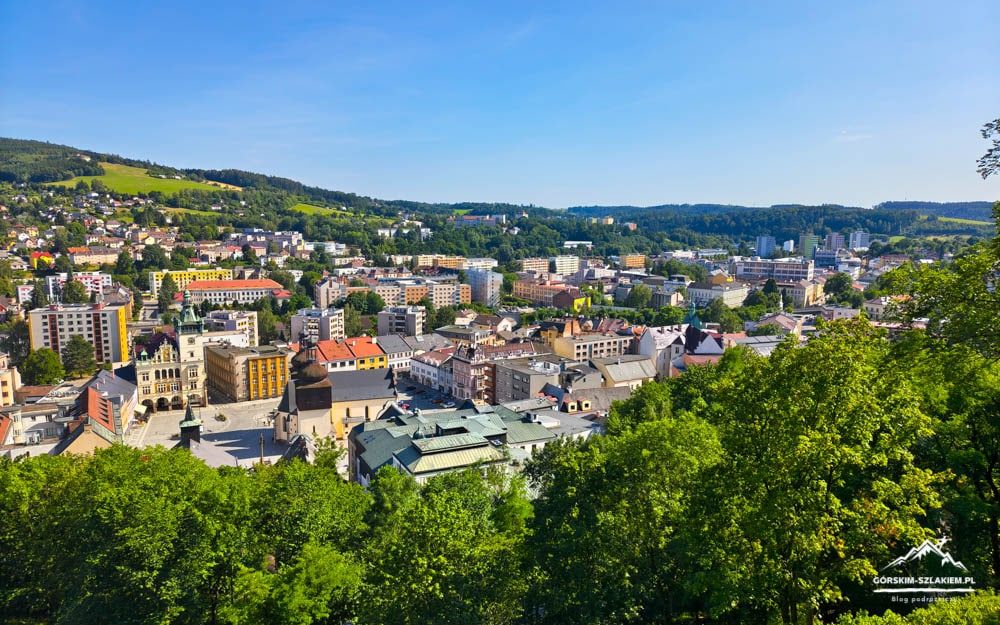 Panoramic view of a town in a valley with colorful buildings, a church, and distant hills, framed by green trees in the foreground.