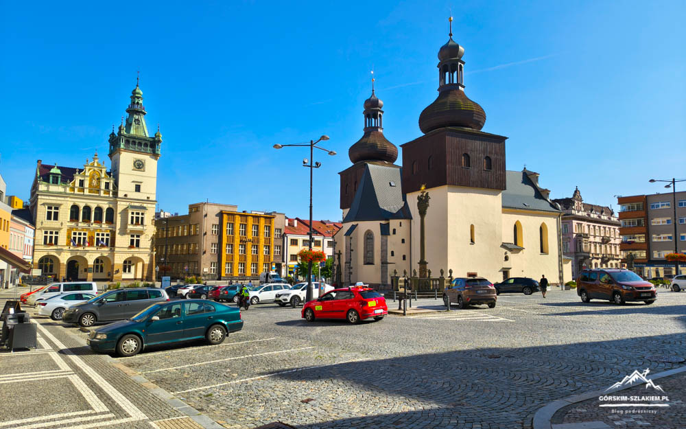 Square in front of a historic church with two dark onion domes; parked cars and colorful buildings under a clear blue sky.