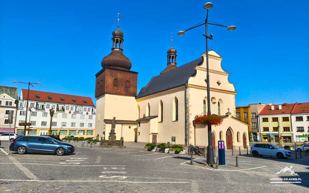 Historic church with a tall wooden bell tower in a sunny town square, parked cars nearby.