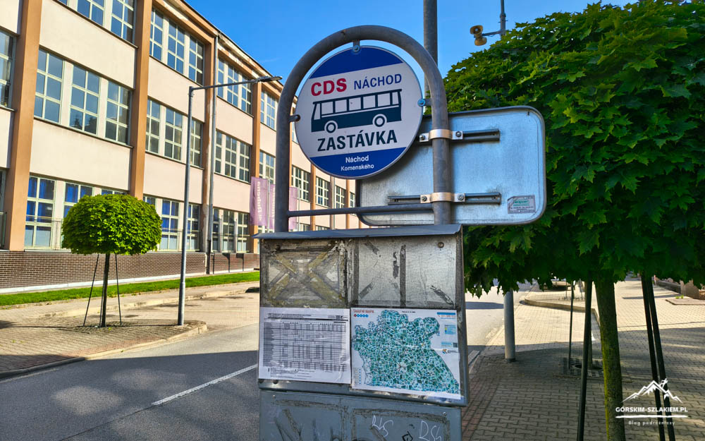 Bus stop sign for CDS Náchod with a bus icon, timetable and map panels, in front of a school with trees and blue sky