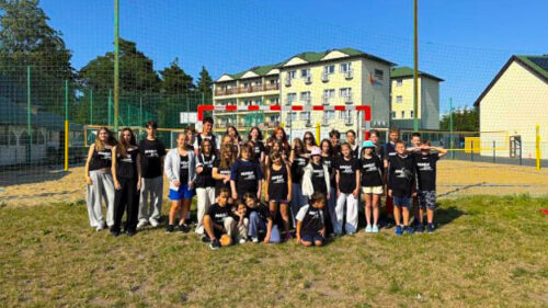 Group of about 30 children and teens in black shirts posing on a grassy field in front of a fenced sports court and residential buildings.