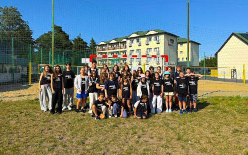 Group of about 30 children and teens in black shirts posing on a grassy field in front of a fenced sports court and residential buildings.