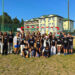Group of about 30 children and teens in black shirts posing on a grassy field in front of a fenced sports court and residential buildings.