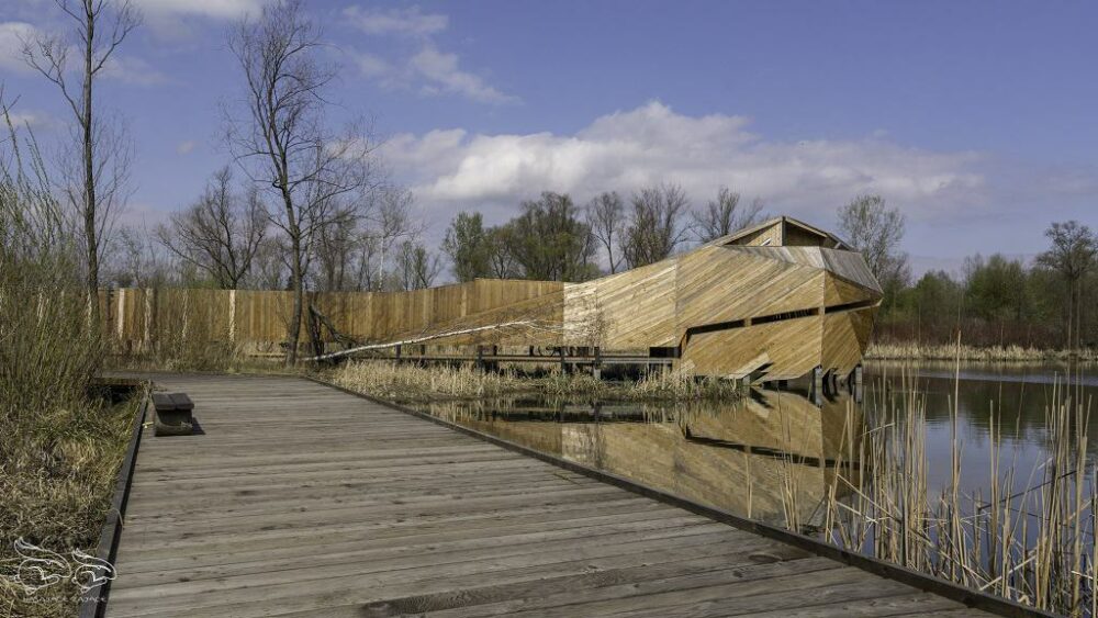 Long wooden boardwalk beside a calm lake leading to an angular wooden structure reflecting in the water.