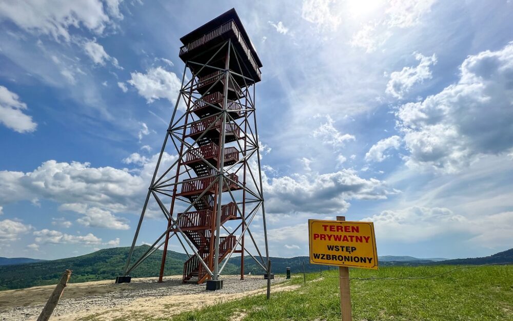 Tall metal observation tower with red spiral stairs on a hill under a blue, cloudy sky; a yellow private-area sign in Polish reading 'Teren prywatny, wstęp wzbroniony' nearby