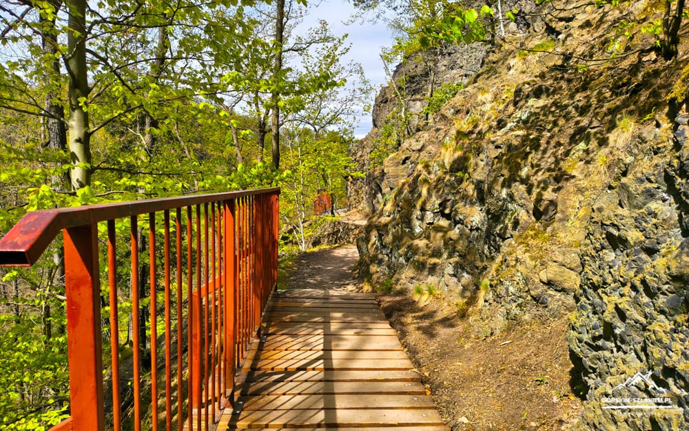 Wooden boardwalk with an orange railing along a rocky cliff, surrounded by a green forest on a sunny day.