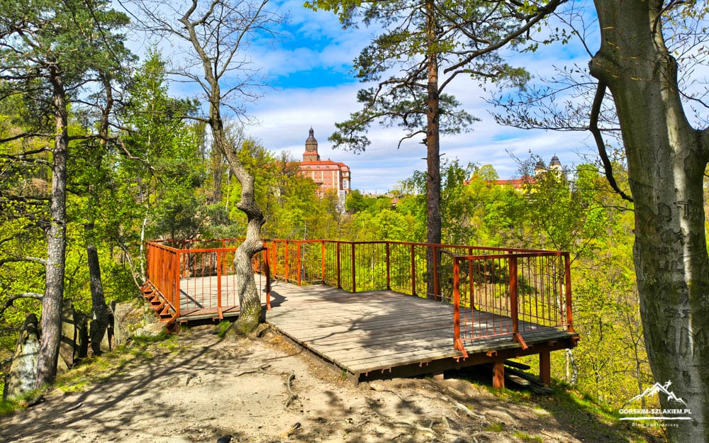 Wooden viewing deck with orange railings in a forest, overlooking distant buildings under a blue sky.