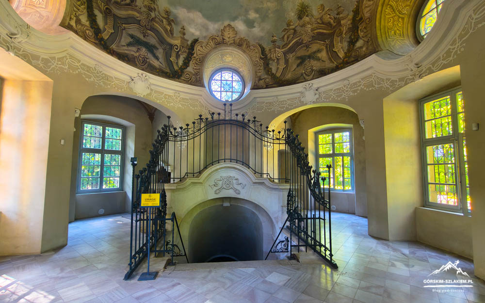Historic interior with a circular black wrought-iron staircase wrapping around a central opening, beneath a frescoed domed ceiling and arched windows.