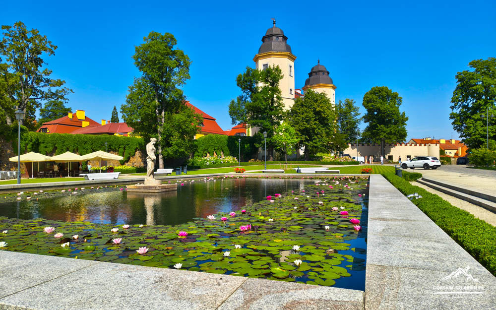 Lily-filled pond with a central statue, surrounded by formal garden paths and trees, with historic towered buildings in the background.