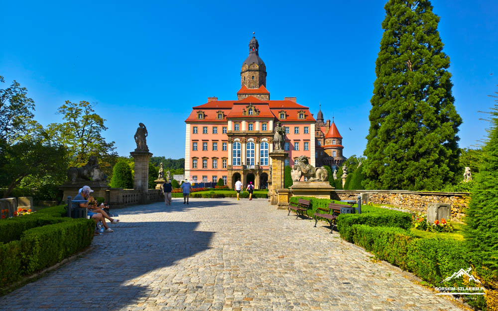 Pink castle with a red roof and tall central tower rises over a cobblestone path lined with statues and hedges in a formal garden. (informative)