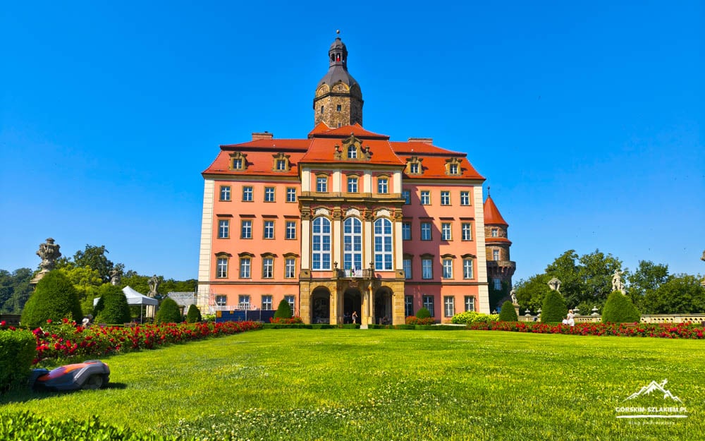 Elegant pink castle with a red roof and central clock/tower, surrounded by manicured gardens and flowering hedges against a bright blue sky.