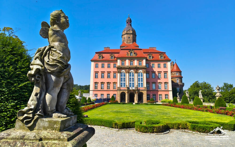 Pink European castle with red roof and central tower, set in formal gardens and gravel path, viewed from a statue-filled foreground