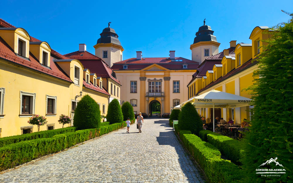 Yellow castle-like complex with a cobblestone courtyard leading to a central arched entrance and a cafe on the right under umbrellas on a sunny day.