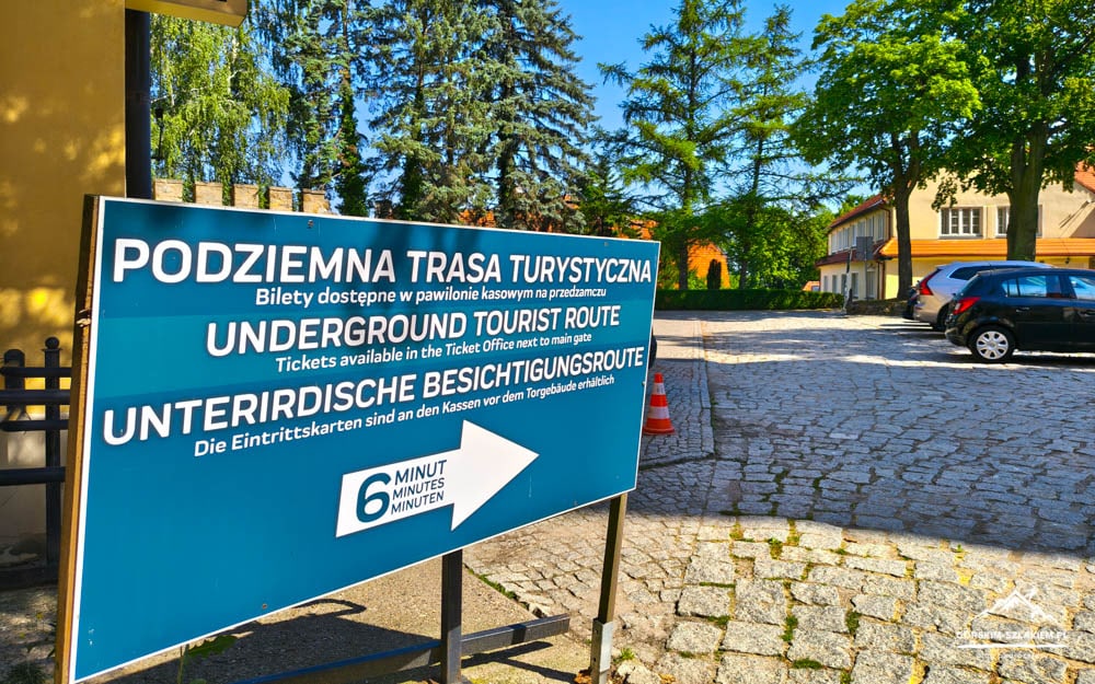 Blue multilingual sign advertising an underground tourist route with a white arrow, on a cobblestone lot beside parked cars.