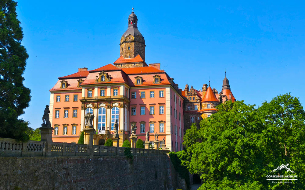 Pink castle with red-tiled roofs and a tall central tower, statues along a stone terrace, against a bright blue sky.