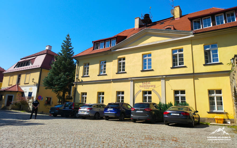 Yellow hotel building with a curved entrance and parked cars along a cobblestone street on a sunny day, blue sky above.