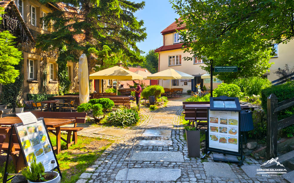 Sunny outdoor cafe courtyard with wooden tables, beige umbrellas, and lush greenery along a stone path between old buildings.