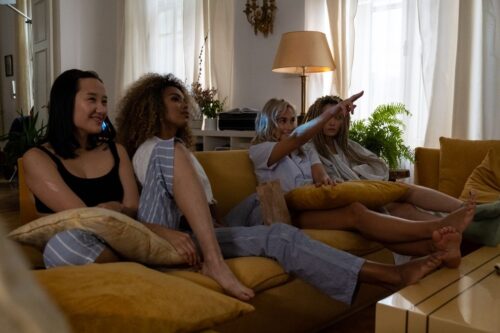 Four women lounging on a mustard-yellow sofa in a bright living room, one pointing at a screen while others smile and chat nearby.