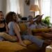 Four women lounging on a mustard-yellow sofa in a bright living room, one pointing at a screen while others smile and chat nearby.